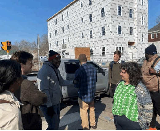 seven people talk outside as a pickup truck drives by in the background